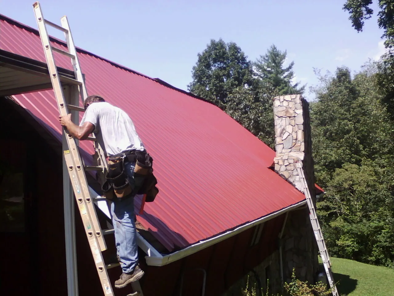 Roofer on a ladder beside a red metal roof and stone chimney — Soffit & Fascia Repair by Roofing Brothers in Edgewater